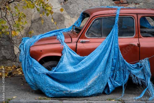 Retro old abandoned red car with broken glass covered with torn blue awning to prevent metal corrosion. Vintage small vehicle forgotten by owner stands on autumn street. unwanted discarded items photo
