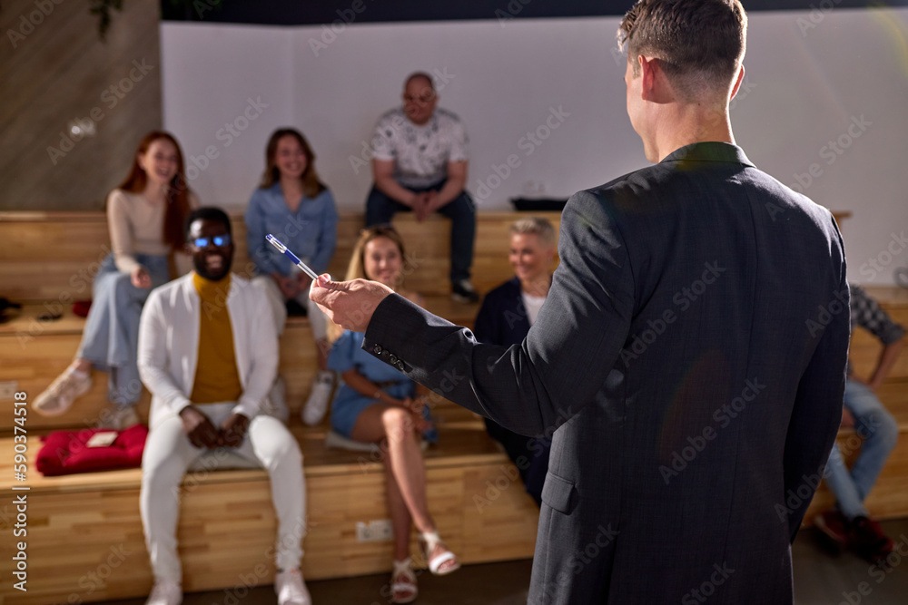 Rear View On Male speaker giving talk in conference hall Pointing With Pen At Side at business event. Audience at the conference hall. Business and Entrepreneurship concept. Male in formal wear Rear View On Male speaker giving talk in conference hall Pointing With Pen At Side at business event. Audience at the conference hall. Business and Entrepreneurship concept. Male in formal wear