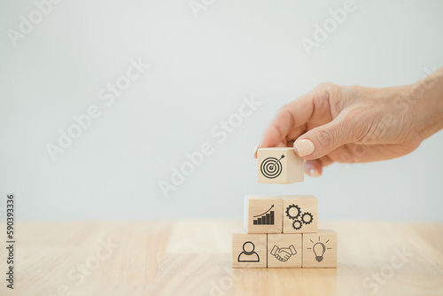 Hand putting wood cube block on top pyramid on table with goal icon on wood cube block, With the concept of a thriving business going for success.