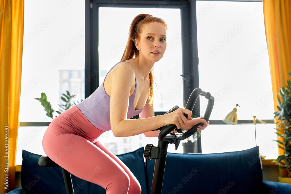 Fototapeta premium Home fitness workout caucasian redhead woman training on stationary bike indoors, engaged in workoud sport. Exercise training for healthy body strong athletic, fit active lady in sportswear