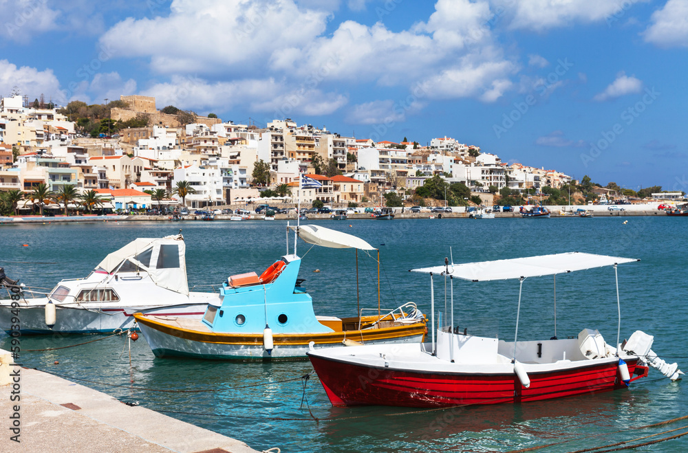 Greece. View of Sitia town from Marina with traditional Greek pleasure bright boats against backdrop of blue sea bay and beautiful white houses on hill topped by ancient Kazarma fortress on sunny day