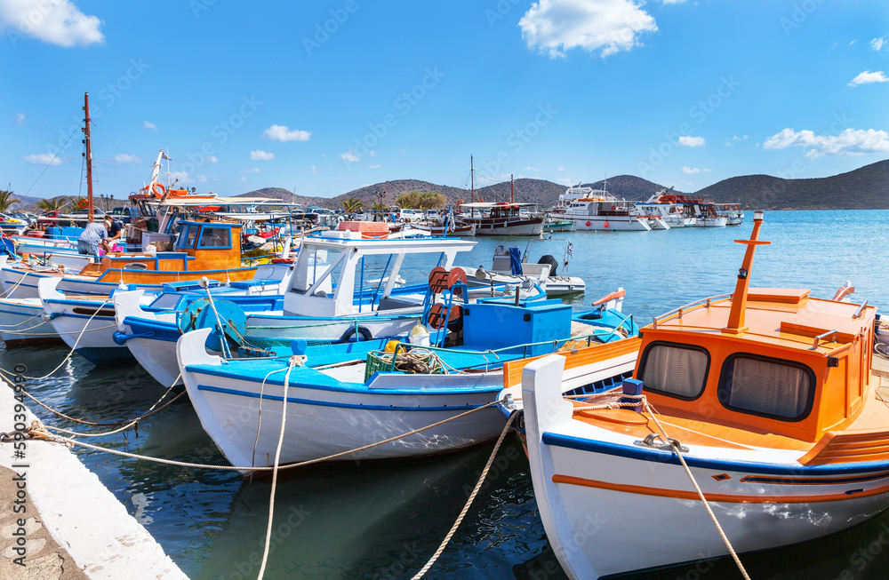 Greece. Crete. Scenic view of Elounda Marina with traditional Greek bright fishing boats on the pier against the backdrop of Mirabello bay on sunny summer day. Seaside recreation and summer travel