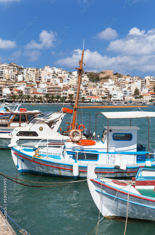 Greece. Beautiful seascape of Marina Sitia with traditional Greek fishing boats against the backdrop of Sitia town with white houses on hill topped by ancient fortress of Kazarma on sunny summer day