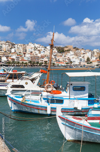 Greece. Beautiful seascape of Marina Sitia with traditional Greek fishing boats against the backdrop of Sitia town with white houses on hill topped by ancient fortress of Kazarma on sunny summer day