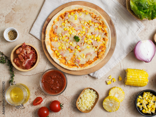 Top view shot of delicious tasty juicy cheesy corn and ham Italian pizza placed on wooden board on party table around with other ingredients, sauce, olive oil, bacon, tomatoes, cheese and vegetables