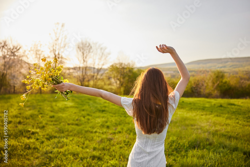 photo from the back of a red-haired woman raising her hands in happiness with a bouquet of flowers in the rays of the setting sun