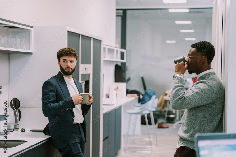 Fototapeta premium Black male person taking a sip of tea while his white colleague is having a conversation with someone. They are in the office kitchen