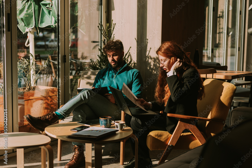 Young business partners working remotely from the coffee bar on a new project. Man is writing in the note book while his female coworker is working on the lap top