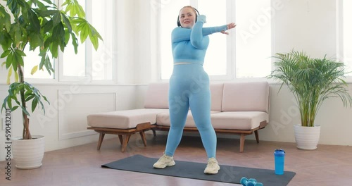 young beautiful overweight happy woman standing on yoga mat and warming up arms. Exercising at home. red-haired woman listening to music while doing sport, slow motion
