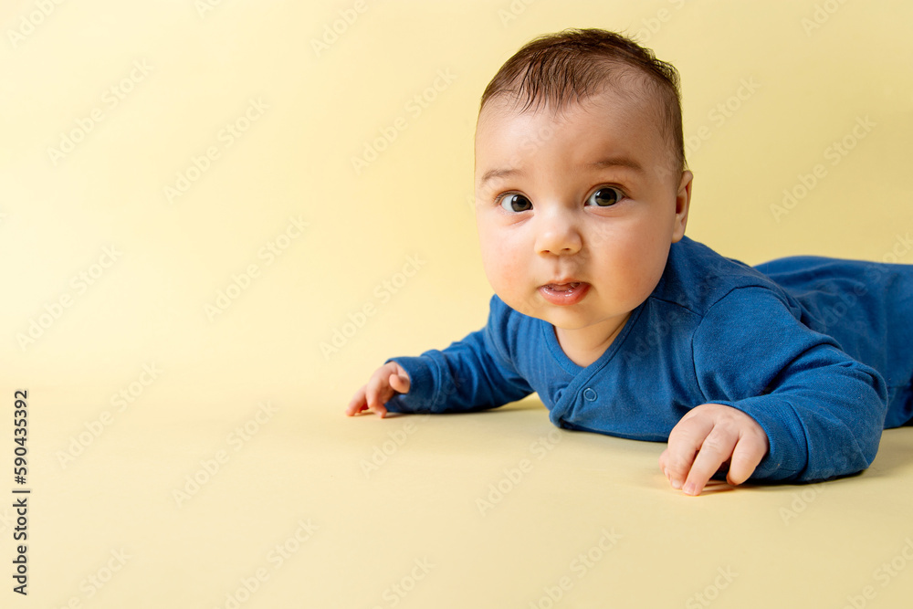 A beautiful little baby in a blue bodysuit, lies on the stomach on a yellow background. Concept, mockup, blank, copy space. A beautiful little baby in a blue bodysuit, lies on the stomach on a yellow background. Concept, mockup, blank, copy space.