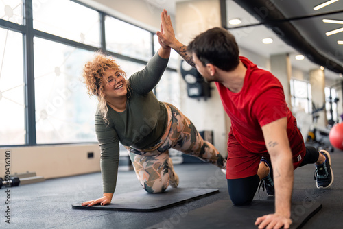 Male personal trainer giving high five support and motivation to smiling young woman fitness client while doing push-ups on exercise mat in gym near panoramic window during individual training.