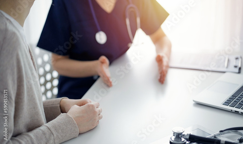 Doctor and patient sitting at the table in clinic while discussing something. The focus is on female patient's hands, close up. Medicine concept