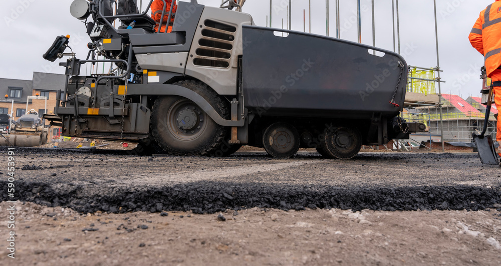 Asphalt paver filled with hot tarmac laying new road surface on new residential housing development site and roadworker operator in orange hi-viz next to it Asphalt paver filled with hot tarmac laying new road surface on new residential housing development site and roadworker operator in orange hi-viz next to it