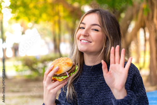 Young pretty Romanian woman holding a burger at outdoors saluting with hand with happy expression