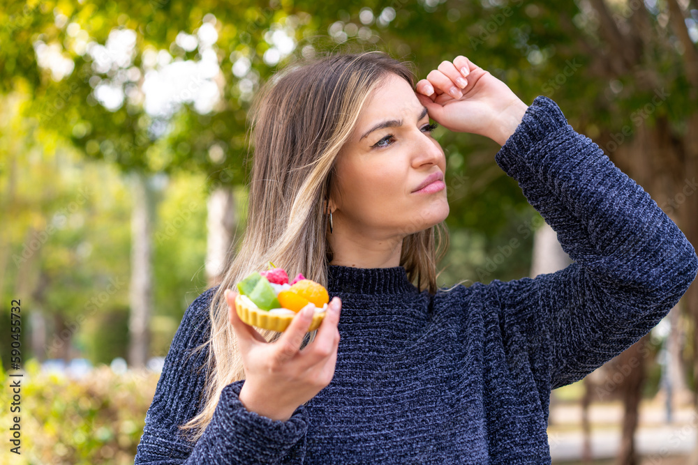 Young pretty Romanian woman holding a tartlet at outdoors having doubts and with confuse face expression Young pretty Romanian woman holding a tartlet at outdoors having doubts and with confuse face expression