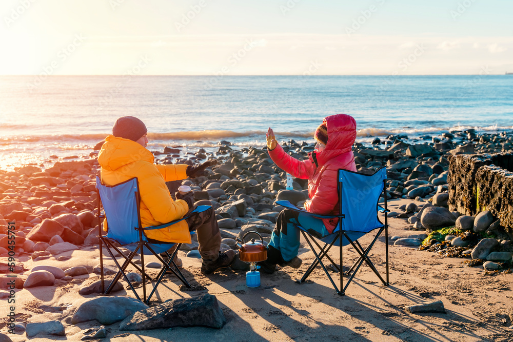 Two people on beach chairs together on winter beach, sunny, blue sky horizon. British cold winter. Local tourism concept.