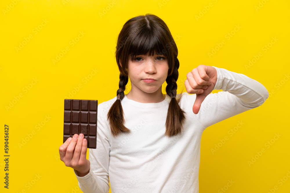 Little caucasian girl isolated on yellow background taking a chocolate tablet making bad signal Little caucasian girl isolated on yellow background taking a chocolate tablet making bad signal