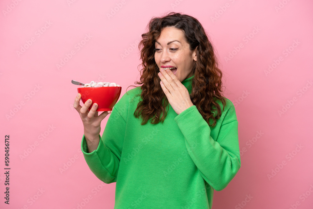 Young caucasian woman holding a bowl of cereals isolated on pink background with surprise and shocked facial expression