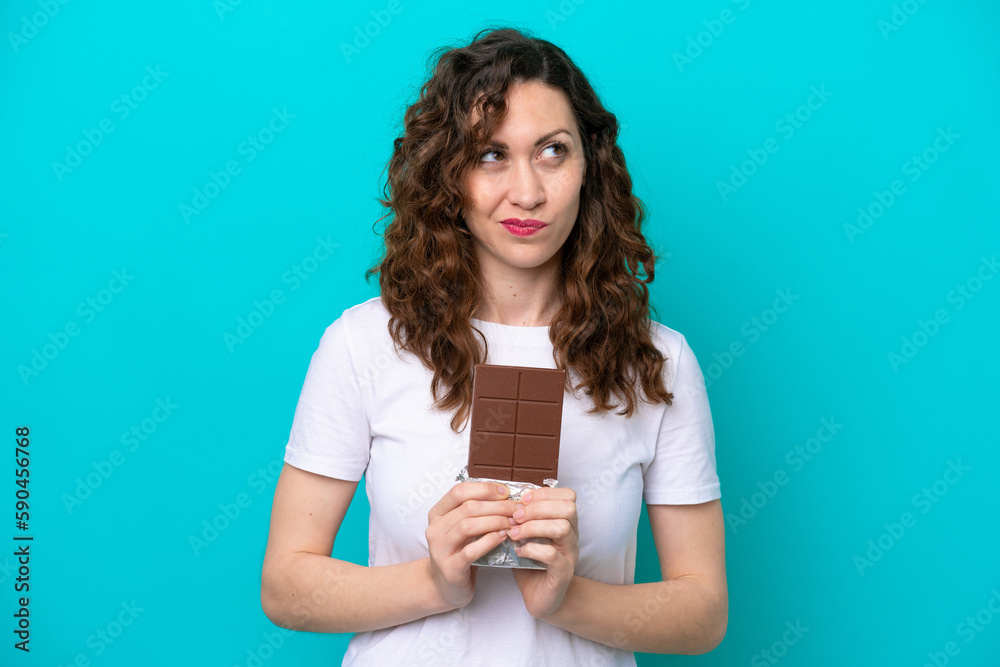 Young caucasian woman isolated on blue background taking a chocolate tablet and having doubts Young caucasian woman isolated on blue background taking a chocolate tablet and having doubts