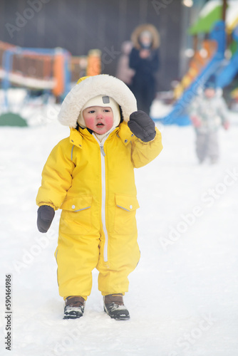Portrait of a toddler baby in warm yellow clothes on a winter playground. Happy baby boy in snowsuit standing in the snow in kindergarten. Kid age one year eight months