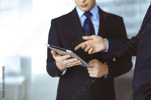 Business people use a tablet computer for discussion of their new project, standing in a modern office. Unknown businessman or male entrepreneur with a colleague at workplace.