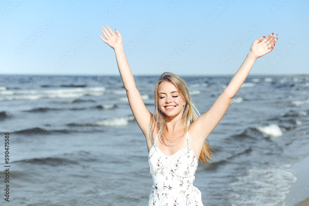 Happy blonde beautiful woman on the ocean beach standing in a white summer dress, raising hands Happy blonde beautiful woman on the ocean beach standing in a white summer dress, raising hands