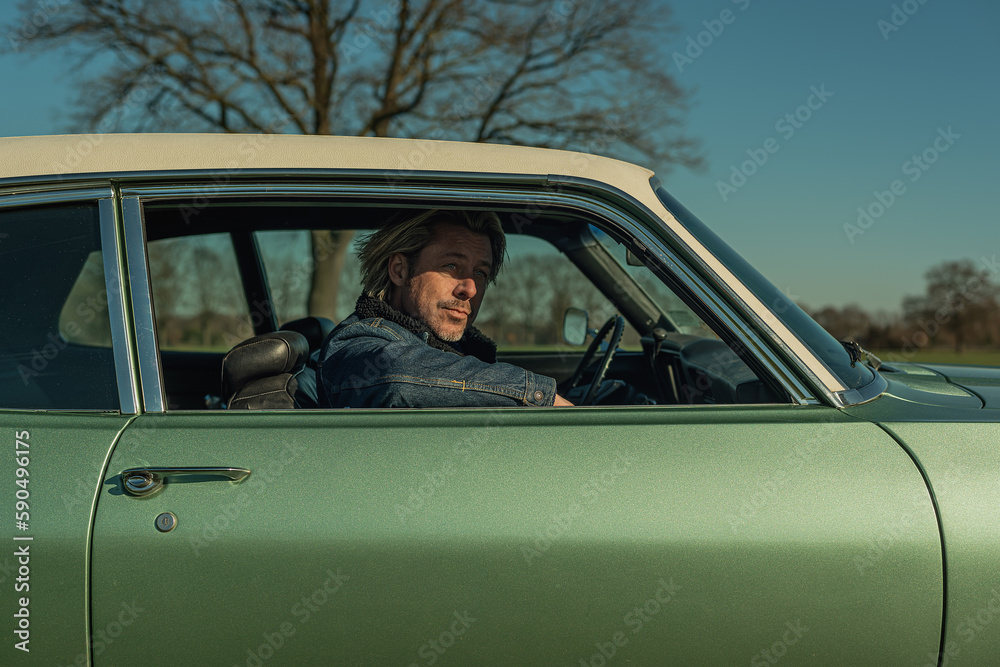 Man with blond hair in jeans jacket sits on passenger side of a vintage american muscle car looking out the window.