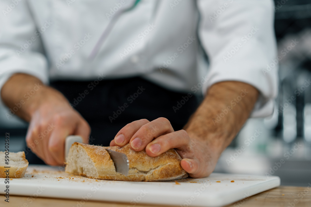 Professional modern restaurant kitchen chef cutting freshly baked ready bread from toast oven with knife close up