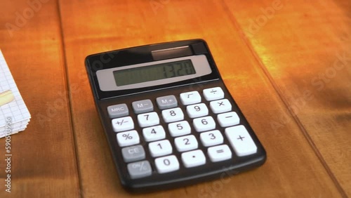 A calculator and a notebook on a brown wooden table under table lamp with warm light, slow motion video.