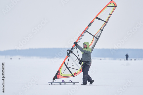 A middle-aged man, a snowsurfer, rolls out a sailboard onto a snow-covered frozen lake. Preparing for snowsurfing on a cloudy winter day.