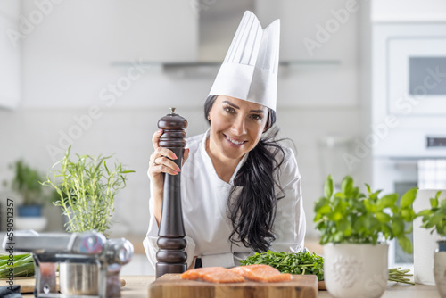 Female chef in toque blanche, white hat, and uniform holds a pepper grinder during fish and veg meal preparation