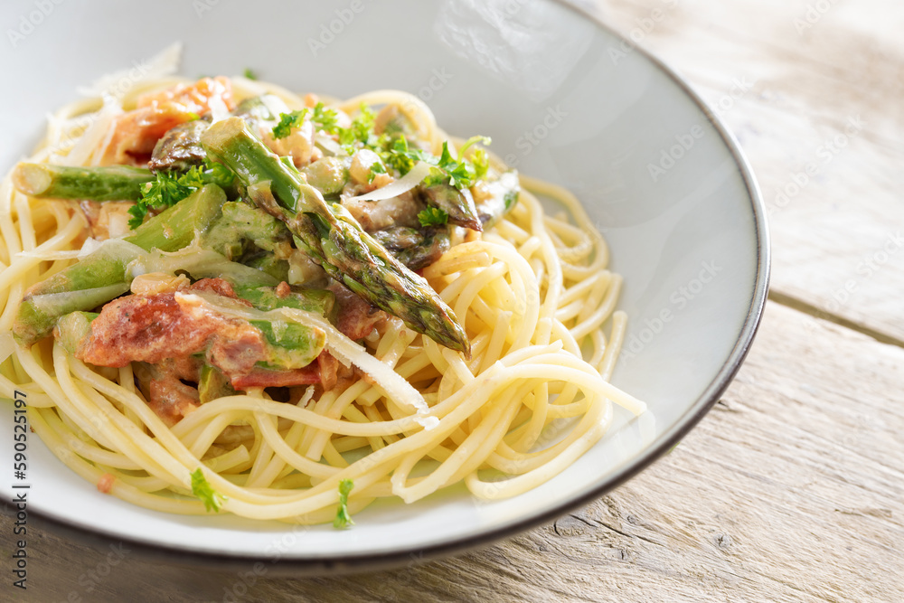 Green asparagus in tomato sauce on spaghetti with parmesan and parsley garnish in a light gray plate on a rustic wooden table, copy space, selected focus