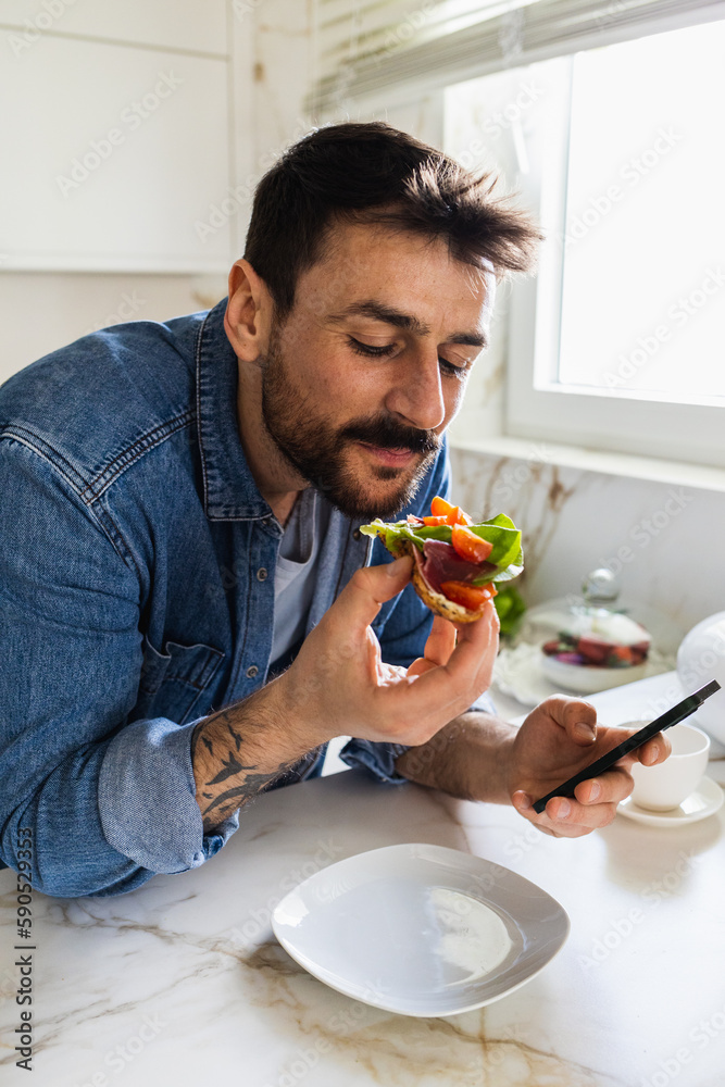 Close-up shot of a handsome young man eating a sandwich for breakfast, while holding a phone.
