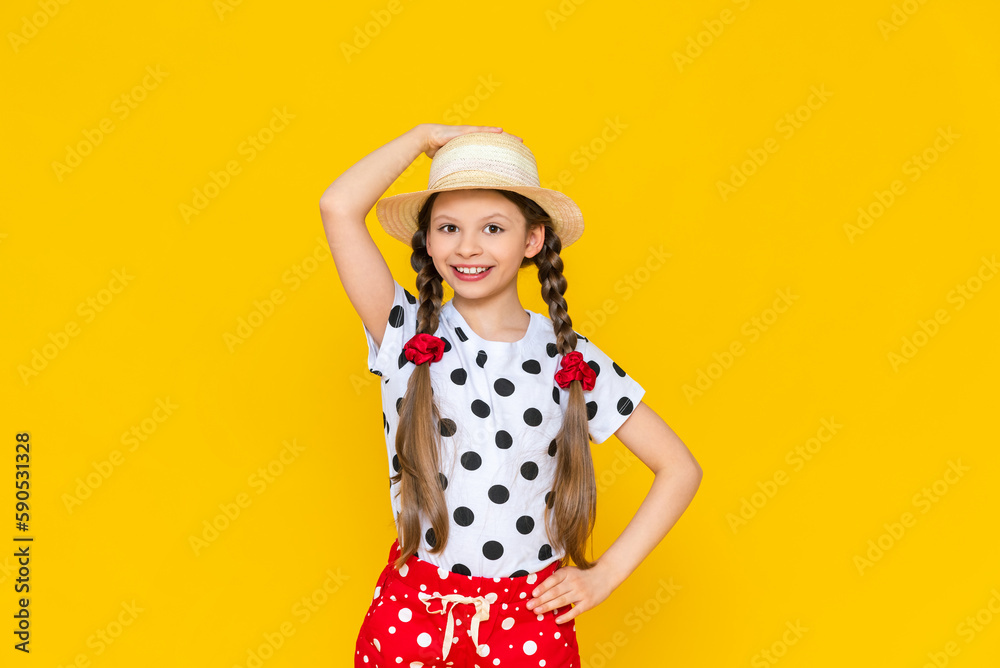 A happy little girl is holding onto a straw hat and smiling broadly, enjoying the bright summer. A teenage girl in polka dot clothes and two pigtails on a yellow isolated background.