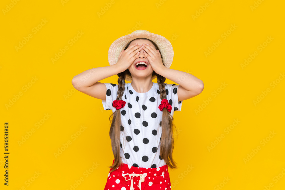 A little girl in a straw hat has closed her eyes with her palms and is smiling broadly, rejoicing in the summer. A happy child in polka dot clothes on a yellow isolated background. A little girl in a straw hat has closed her eyes with her palms and is smiling broadly, rejoicing in the summer. A happy child in polka dot clothes on a yellow isolated background.