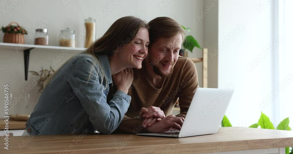 Cheerful happy millennial husband and wife shopping online, booking trip, hotel for vacation, using app on laptop computer in home kitchen for Internet communication Cheerful happy millennial husband and wife shopping online, booking trip, hotel for vacation, using app on laptop computer in home kitchen for Internet communication