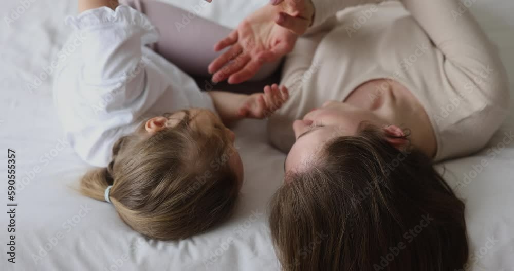 Peaceful young mother and toddler daughter kid relaxing on bed together, holding hands, making arm motion, playing together. Mom calming preschooler little child for sleeping Peaceful young mother and toddler daughter kid relaxing on bed together, holding hands, making arm motion, playing together. Mom calming preschooler little child for sleeping