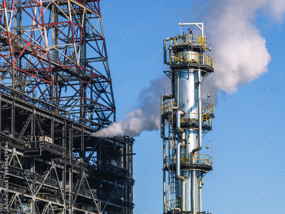 Oil refining plant. Technological equipment for the production of fuel components. White smoke escapes into the atmosphere from the production equipment at the refinery. White smoke and blue sky. Oil refining plant. Technological equipment for the production of fuel components. White smoke escapes into the atmosphere from the production equipment at the refinery. White smoke and blue sky.