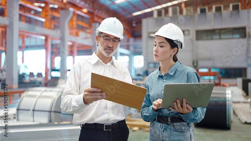Two engineer manager leader and woman assistant holding laptop wearing helmet talking and checking production standing in workplace area at manufacturing factory