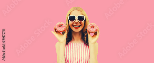 Portrait of happy cheerful laughing young woman with two sweet donuts having fun wearing heart shaped sunglasses on pink background