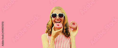 Portrait of happy cheerful laughing young woman with two sweet donuts having fun wearing heart shaped sunglasses on pink background