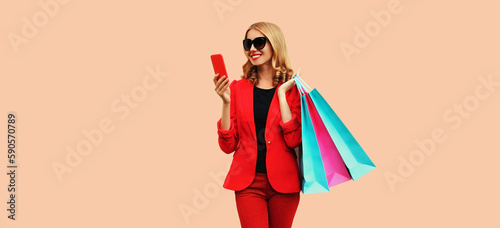 Portrait of happy smiling young woman with smartphone and shopping bags wearing business blazer on pink background