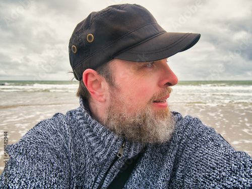 Ein bärtiger, friesisch wirkender Mann mit schwarzer Cap blickt zur Seite während im Hintergrund die Wellen der Nordsee zu sehen sind