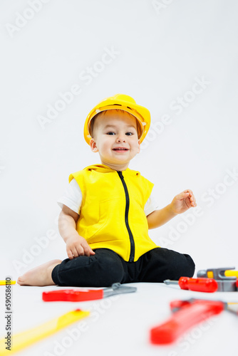 A little boy 2 years old in a builder's suit on a white background. A child in a plastic helmet and plastic tools. Plastic children's toys.