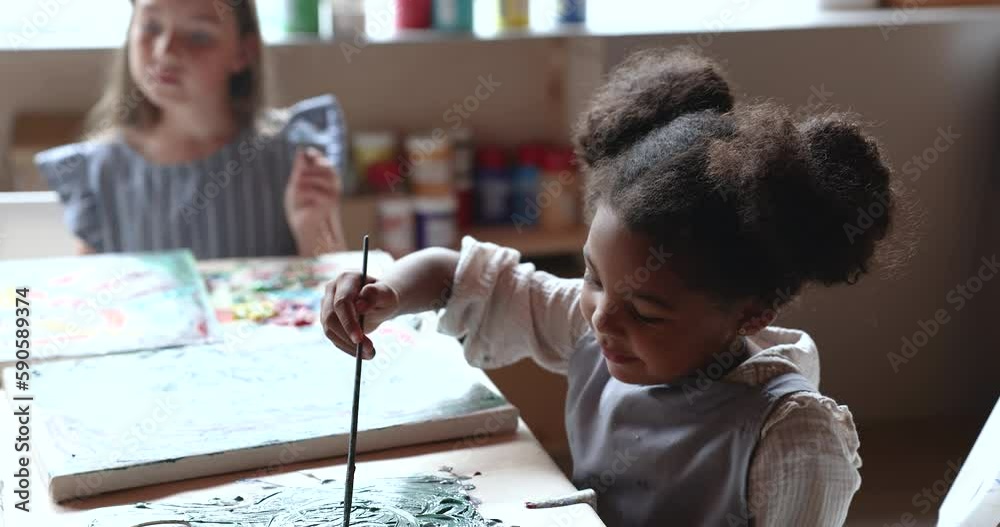 Cheerful cute African kid girl having fun on drawing class, painting on canvas at table, holding paintbrush, laughing, smiling, enjoying creative hobby, childhood, training artistic skills