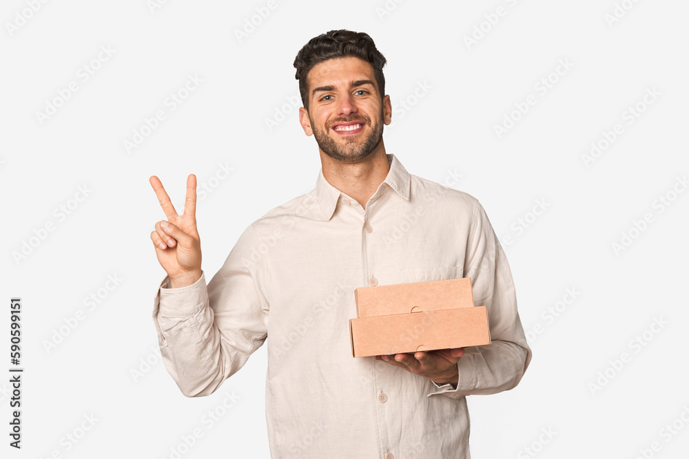 Young man holding a takeaway boxes isolated joyful and carefree showing a peace symbol with fingers. Young man holding a takeaway boxes isolated joyful and carefree showing a peace symbol with fingers.