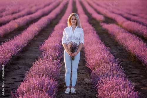Fototapeta Naklejka Na Ścianę i Meble -  Young woman in white shirt, blue jeans stays in rows of purple lavender in field. Millennial girl holding bouquet of flowers in hands at sunset. Allergy concept. Production natural perfumery.