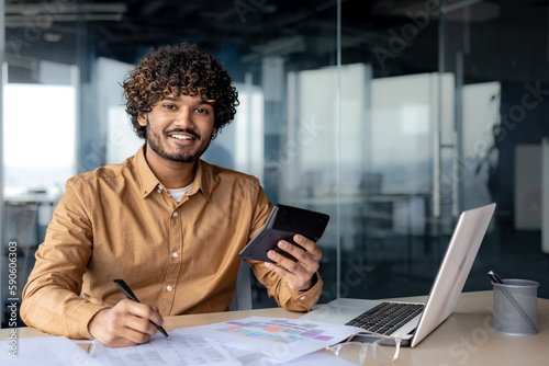 Portrait of young successful financier accountant at work inside office building at workplace, hispanic man smiling and looking at camera, man using calculator in paperwork. photo