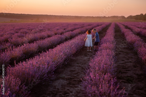 Playful cute boy girl are playing in rows of lavender purple field at sunset. Small couple runs after each other, catches up, holding hands. Cheerful, happy childhood. Travel in countryside. Allergy