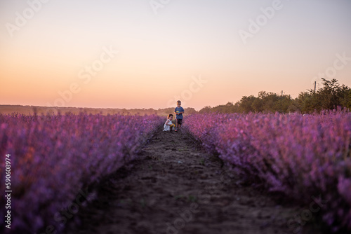 Playful cute boy girl are playing in rows of lavender purple field at sunset. Small couple runs after each other  catches up  holding hands. Cheerful  happy childhood. Travel in countryside. Allergy